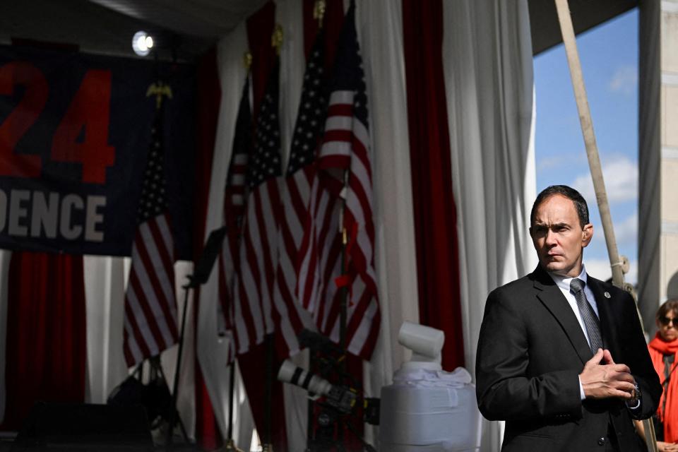 A man in a black suit stands in front of American flags and looks sideways from the stage where Robert F. Kennedy Jr. is slated to speak.