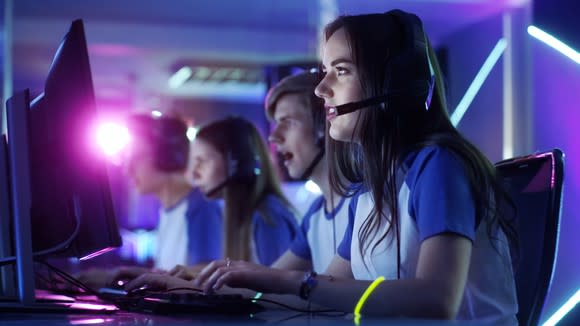 A row of young people wearing team shirts sitting at computers wearing headphones and microphones in a dimly lit room.