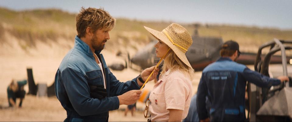 Two actors on set, male in a jacket and female in a hat, conversing with a beach backdrop
