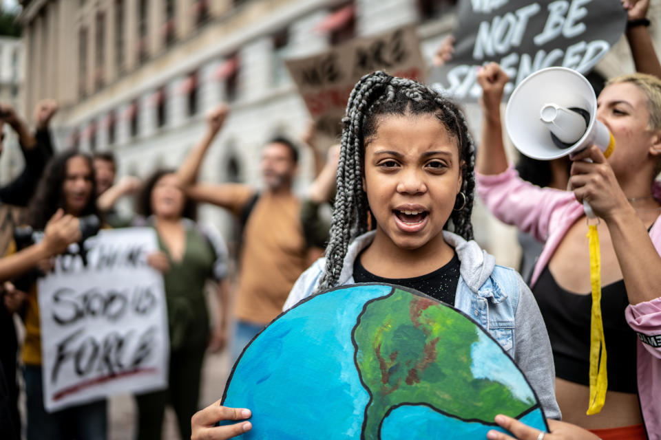 A young girl holds a cardboard Earth while leading a climate change protest with a diverse crowd behind her, one holding a megaphone