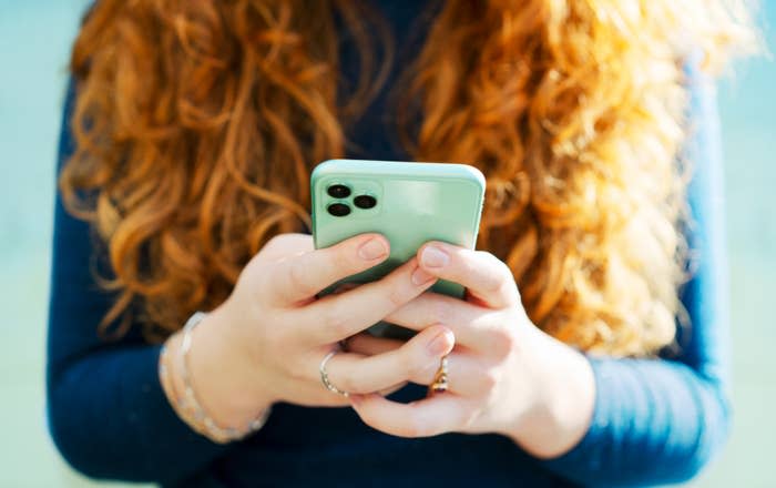 A person with long, curly hair holds a smartphone in both hands, wearing a long-sleeved top and bracelets