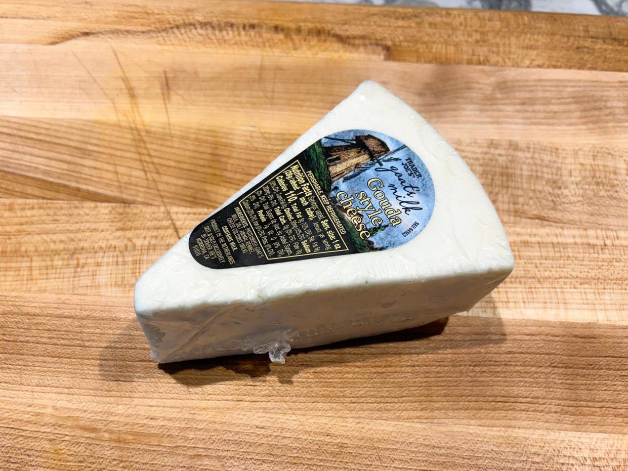White wedge of cheese with a blue label with an illustration of a windmill and text reading "Goat's milk gouda-style cheese" on a wooden cutting board