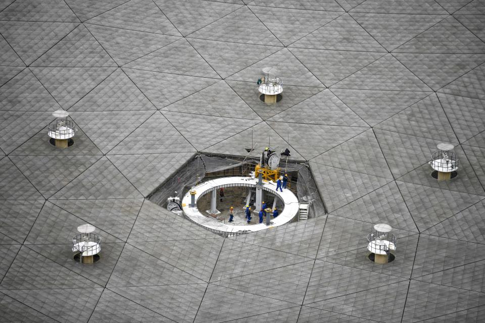 In the bottom of an enormous radio telescope dish, small workers are seen like ants, performing maintenance.