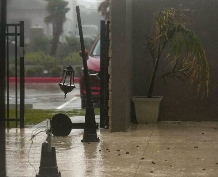 Severe weather causes damaged streetlights and fallen trees in a neighborhood. A car is seen in the background through heavy rain