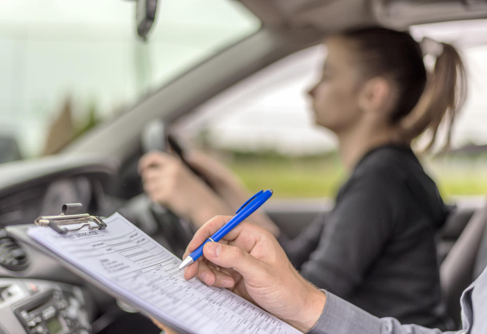 Teenage Girl Taking A Driving Lesson during bright sunny day. Closeup of a instructor man writing on a clipboard and taking notes while sitting next to teen driver.