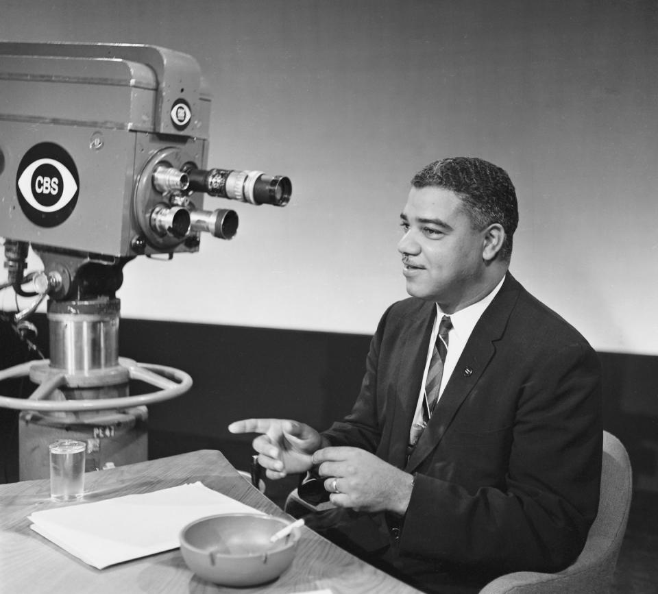 whitney young jr sits at a table and points with one finger, he wears a suit jacket and patterned tie, a cigarette rests in an ash tray in front of him