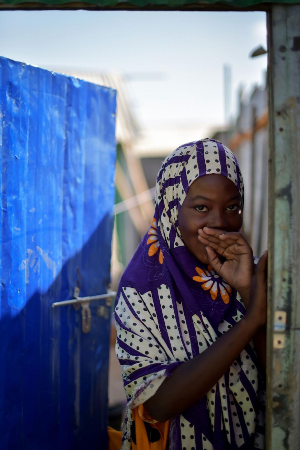 <p>A young Somali girl smiles while standing in the doorway to her home at the Dayniile Persons (IDP) camp in Mogadishu, Somalia, on March 6, 2017. (Photo: Tobin Jones/UN) </p>