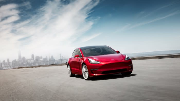 A red Tesla Model 3 with a cityscape in the background.