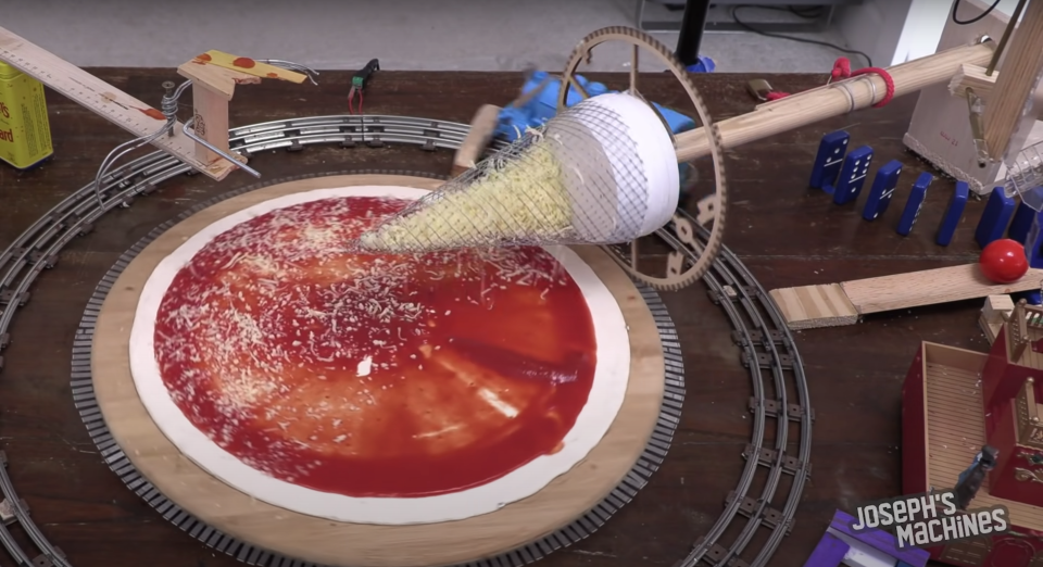 A wacky pizza-making robot uses its cheese-cone to sprinkle cheese on a mid-production pizza.