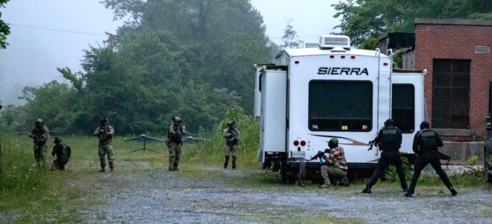 Special operators participating in the Ridge Runner 23-01 exercise advance through an area with members of the opposing force seen hiding behind a trailer. What appears to be two mock drones on stands are seen in the background to the left. <em>Army National Guard </em> Personnel participating in Ridge Runner 23-01 advance through an area with members of the opposing force seen hiding behind a trailer. What appears to be two mock drones on stands are seen ion the background to the left. <em>Army National Guard </em>