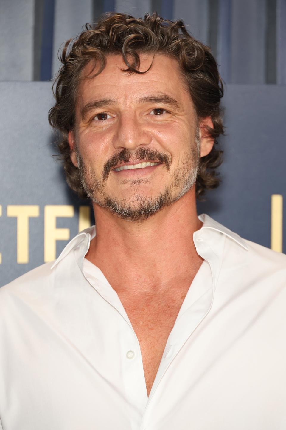 Pedro Pascal wearing a white shirt with an open collar, standing in front of a backdrop with text