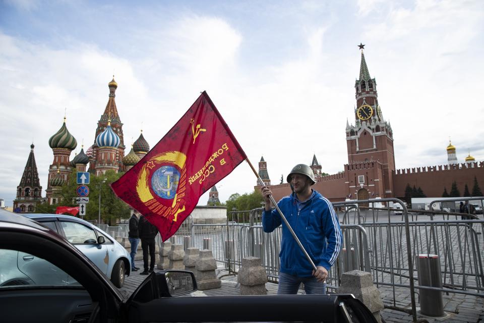 A man poses for a photo with a red flag in front of the closed Red Square during the 75th anniversary of the Nazi defeat in World War II in Moscow, Russia, Saturday, May 9, 2020. Victory Day is Russia's most important secular holiday and this year's observance had been expected to be especially large because it is the 75th anniversary, but the Red Square military parade and a mass procession called The Immortal Regiment were postponed as part of measures to stifle the spread of coronavirus. (AP Photo/Pavel Golovkin)