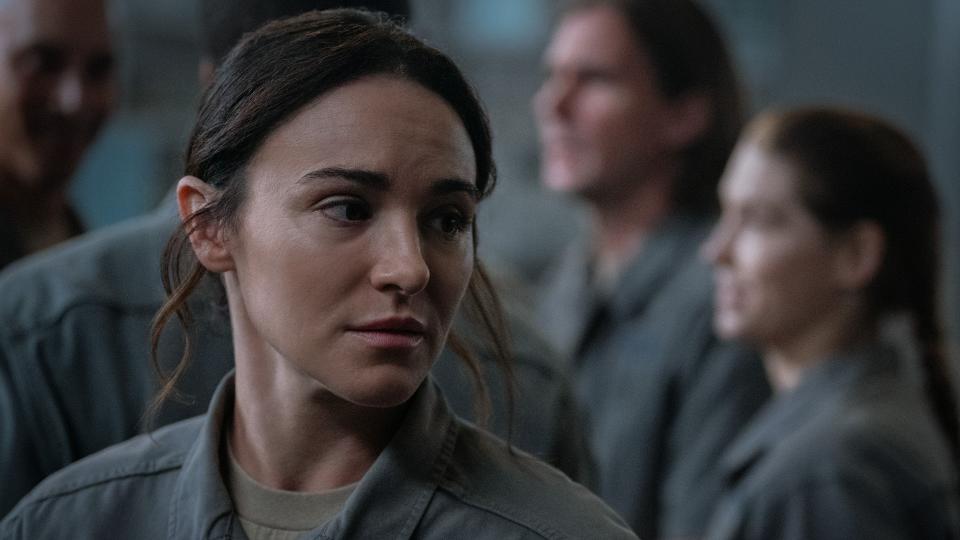 Close up of a worried-looking woman with long dark hair tied up. She is wearing a gray jumpsuit. In the background you can see several other people also wearing the same gray jumpsuit.