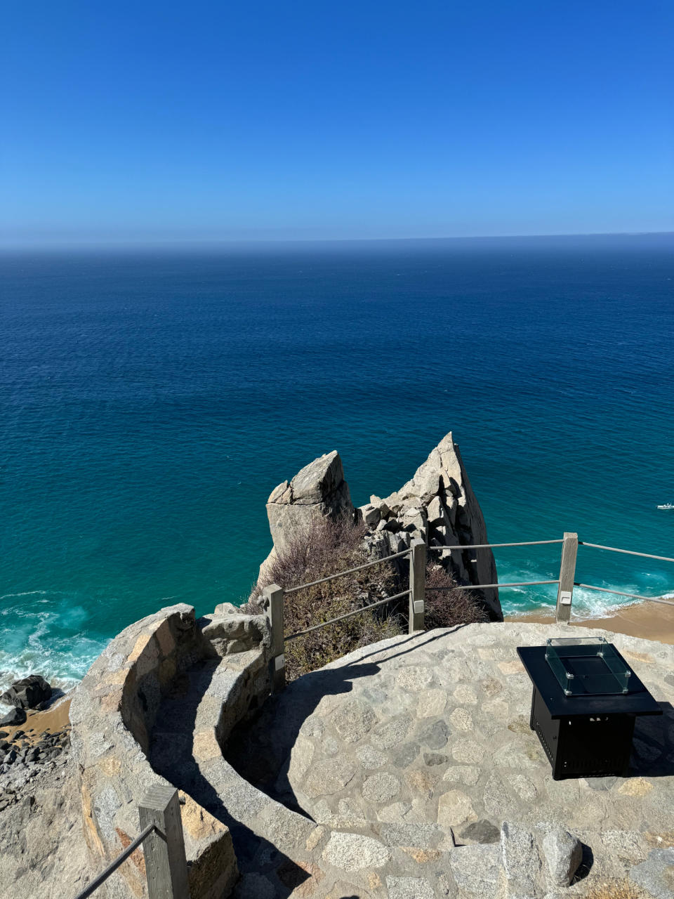 Oceanview from a stone walkway winding around a cliff edge, overlooking a beach and expansive water horizon. No people are present