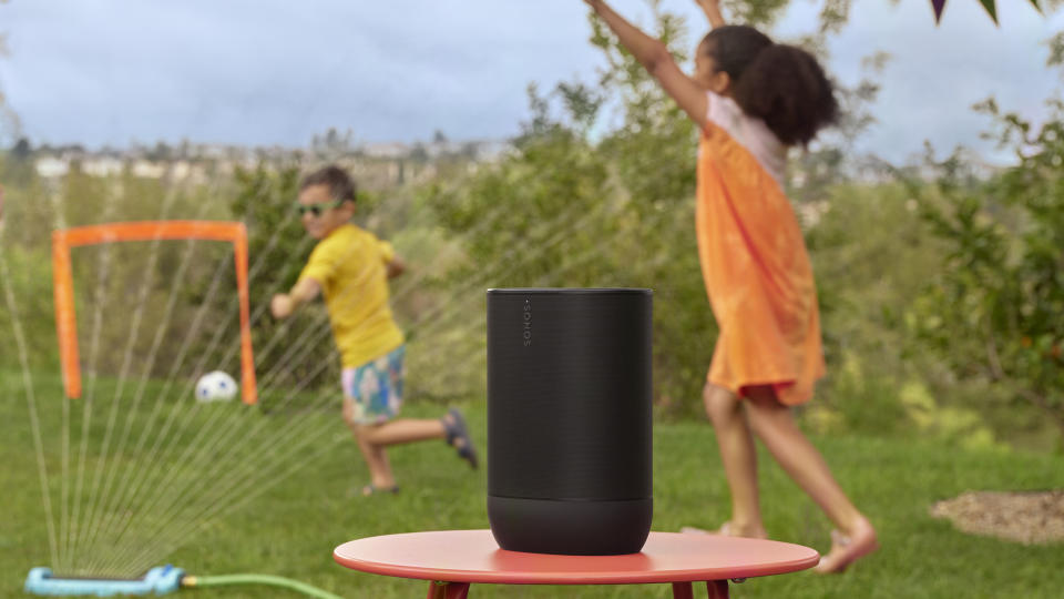 Sonos Move 2 on a table outside, with children playing in the spray from a sprinkler behind it