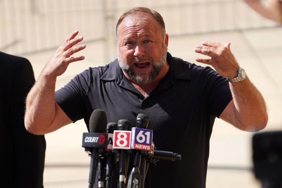 Man gesturing while speaking at a podium with multiple microphones from news outlets