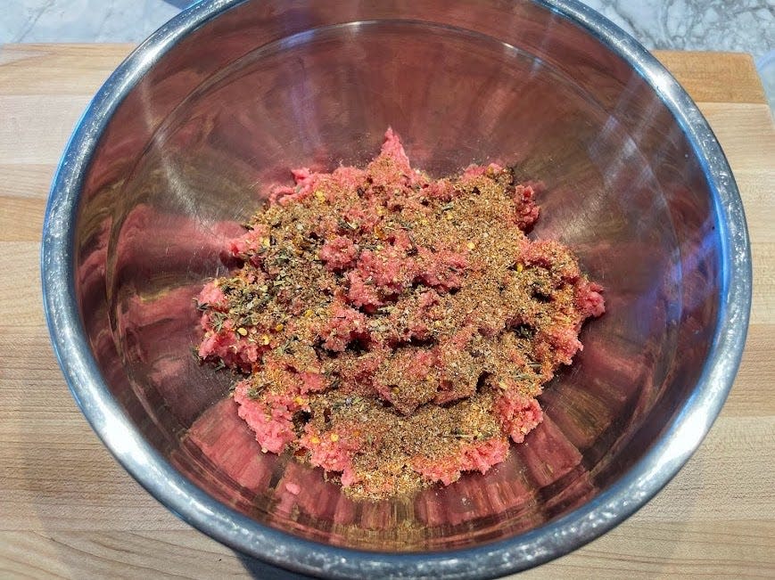 A metal placed on a wooden cutting board filled with ground beef. The beef is sprinkled with spices