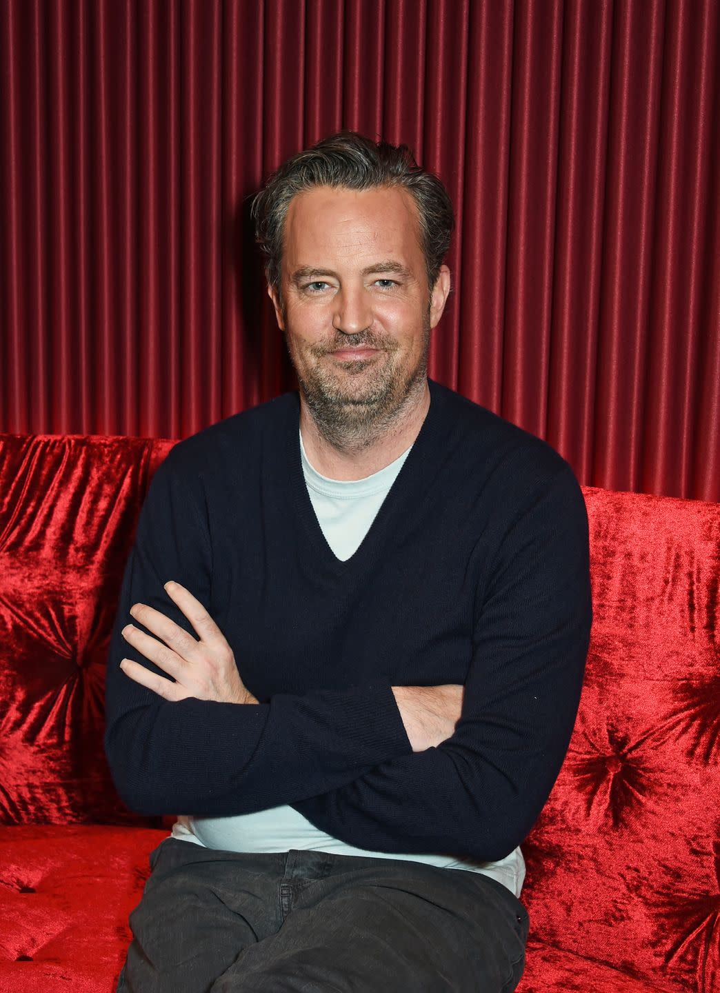matthew perry sits with his arms folded against a red sofa with a matching red curtain behind him