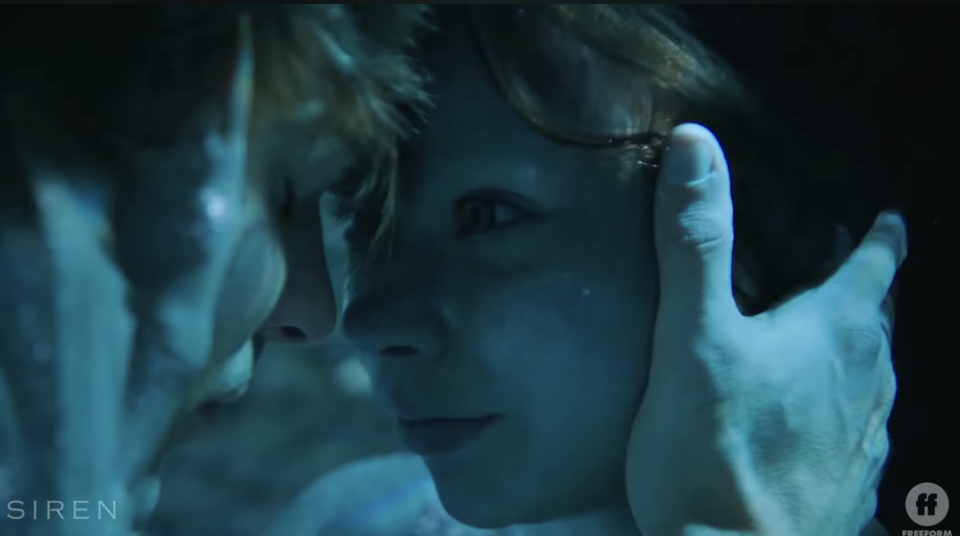 Eline Powell as Ryn in "Siren," holding her lover, Ben, in an embrace in her mermaid form while they're underwater