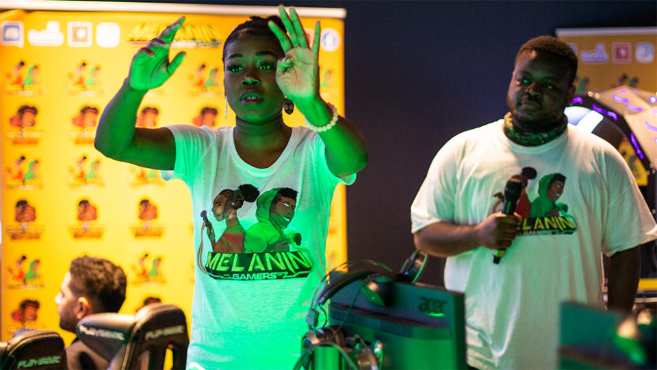 A woman in a white t-shirt - printed with a design of two black characters wearing gaming headsets and the words "Melanin Gamers" - signals to someone off-camera. Behind her, a man wearing an identical shirt and holding a microphone looks on