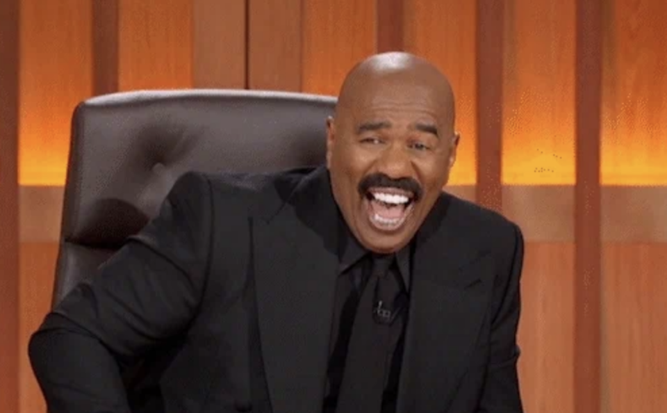 Steve Harvey, wearing a black suit, is seated and laughing in a studio setting