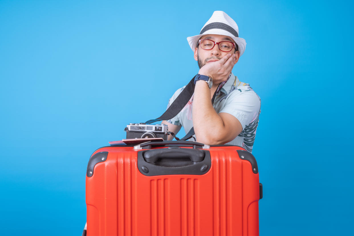 A sighing male tourist leans against a suitcase, with a camera in one hand and his face in the other.