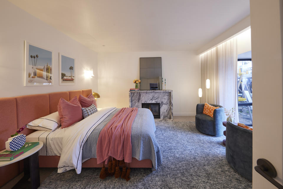 A wide shot of the pink queen sized bed on the left a fireplace on the far wall and two blue chairs on the right. 