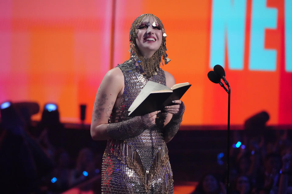 Chappell Roan smiling and holding a book on stage, wearing a shimmering, avant-garde outfit with a facemask, at an award ceremony