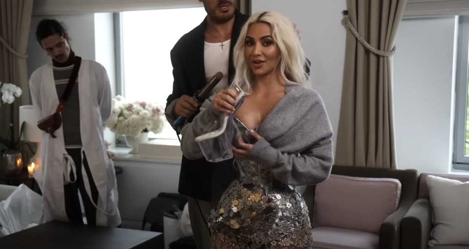 Kim Kardashian in a sparkling dress holding a glass bottle, with two men in the background, one holding a microphone
