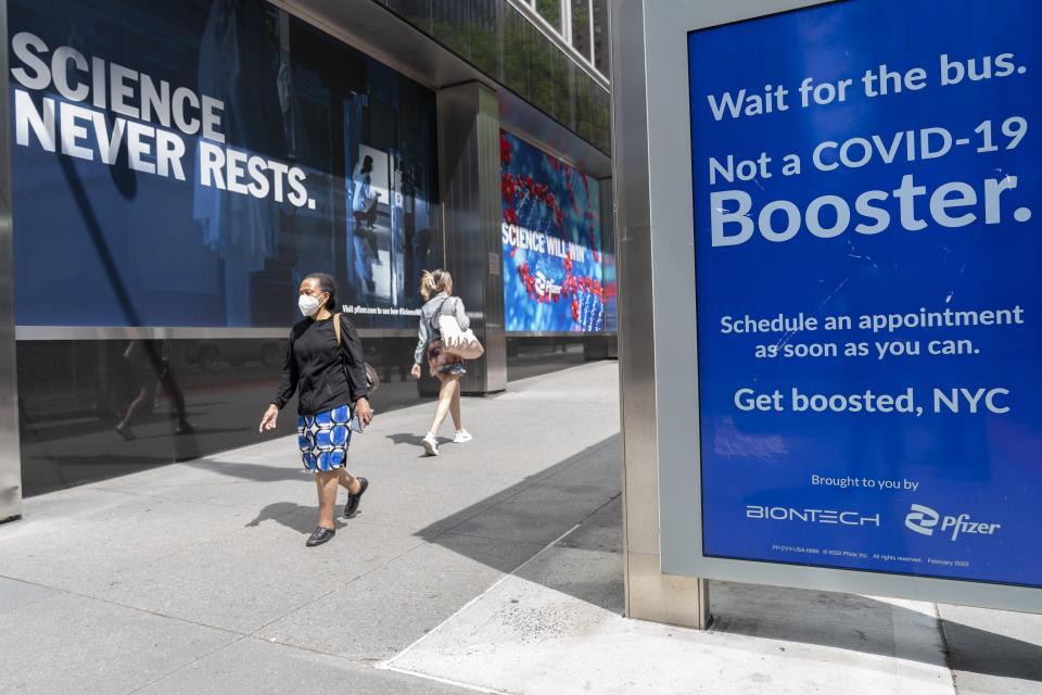 Pedestrians walk past a signs hanging outside Pfizer headquarters in New York and one hanging at a bus stop encouraging the Covid-19 booster, Monday, May 23.