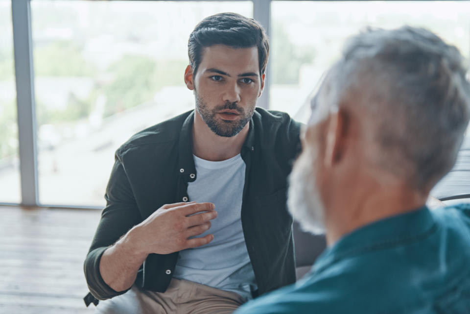 Two men are sitting and having a deep conversation in a brightly lit room with large windows. One man gestures as he speaks