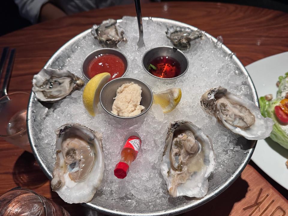 A bowl filled with crushed iced and several oysters, lemon slices, and hot sauce in the bowl. The bowl sits on a wooden table with a salad in the background