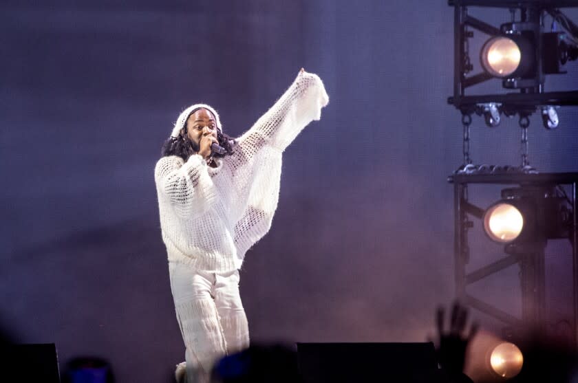 A man rapping into a microphone and raising his arm in an all-white outfit on a stage