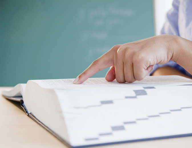 A person's hand points to text in an open book on a desk