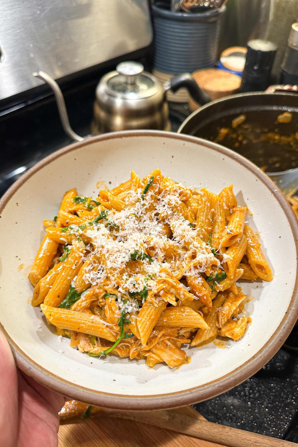 A person is holding a bowl of penne pasta topped with grated cheese; a kitchen stove and utensils are in the background