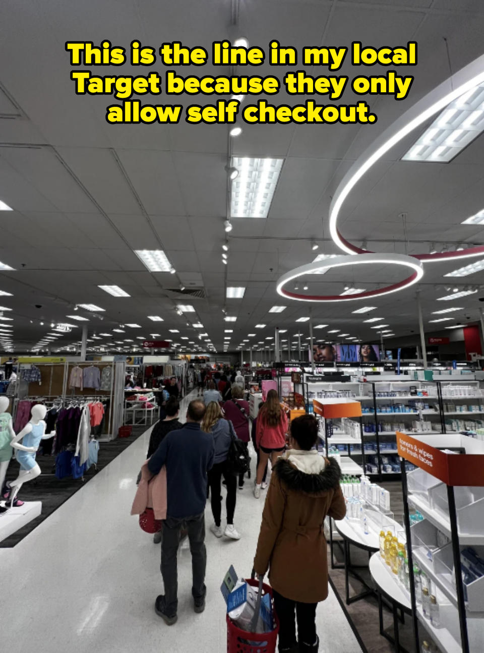 People walk through a busy store aisle lined with various products and clothing sections. The store has a high ceiling with bright lighting