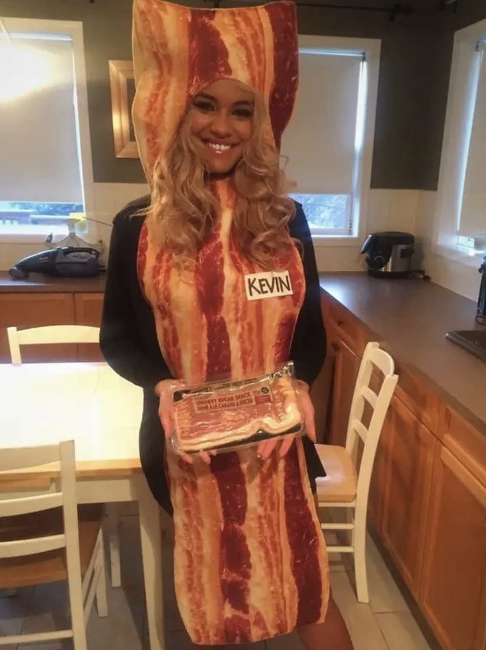 A woman dressed as a strip of bacon, holding bacon, wearing a name tag that says "Kevin"