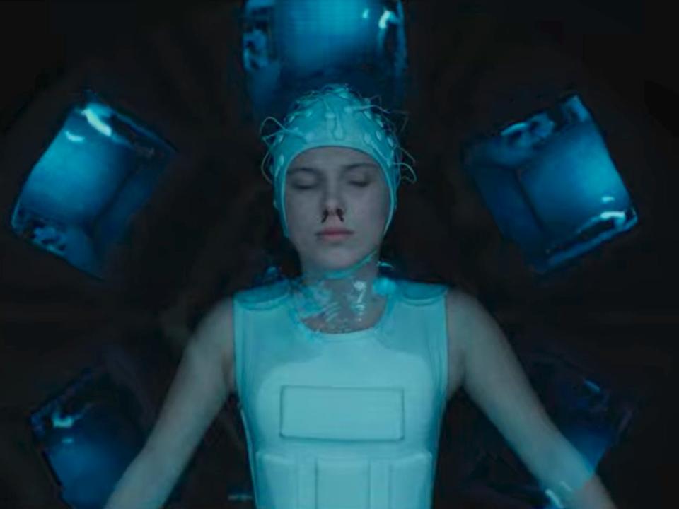 eleven in stranger things floating in a tank, wearing a white vest and a cap on her head with wires protruding from it. her eyes are closed and she has a bloody nose