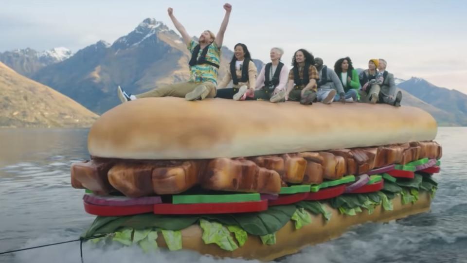 Eight people ride atop a giant replica subway sandwich on a lake surrounded by mountains.