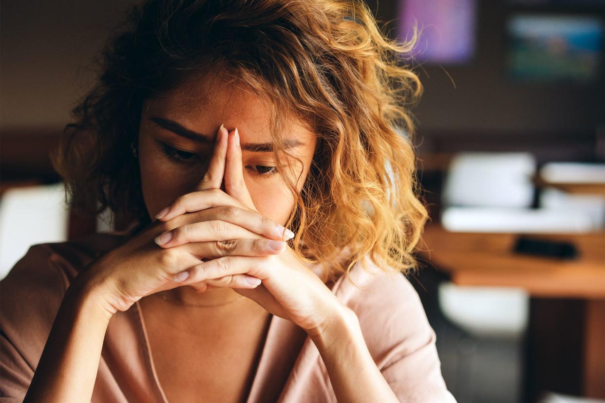 Front-view of very stressed woman's face and shoulders, she is looking towards the bottom-right with her hands arranged on her face for a tension headache, darken room blurred in the background