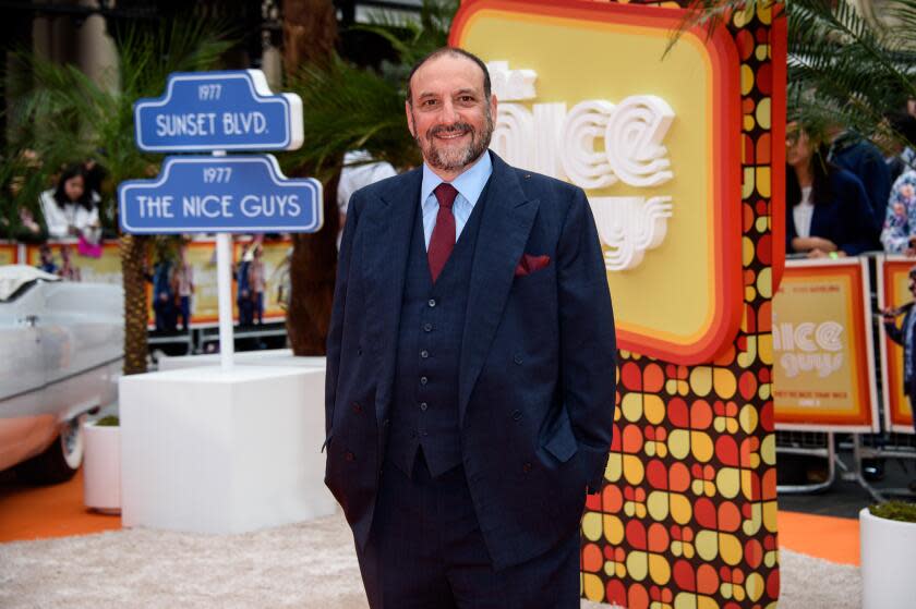 Joel Silver smiles and poses with his hands in his pockets while wearing a blue suit in front of a colorful sign.