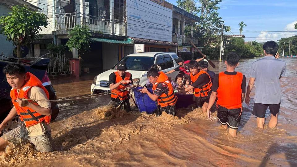 Men in orange life jackets pull along a small boat full of children through waist-high water in a street.