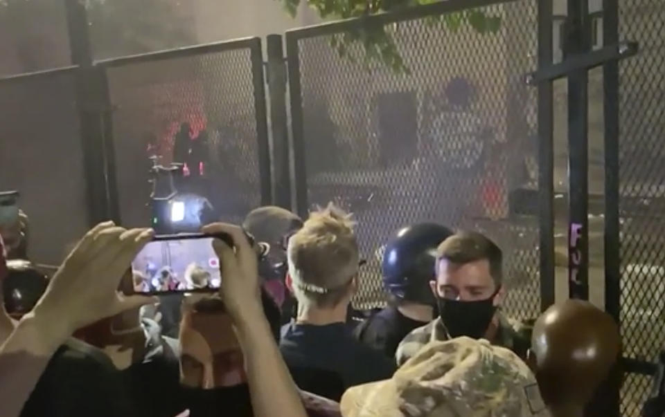 In this image made from video released by Jonathan Maus, Portland Mayor Ted Wheeler, center in black looking away, stands at a fence guarding a federal courthouse as tear gas drifts by early July 23, 2020, in Portland Oregon, during another night of protest against the presence of federal agents sent by President Donald Trump to quell unrest in the city. (Jonathan Maus/BikePortland via AP)