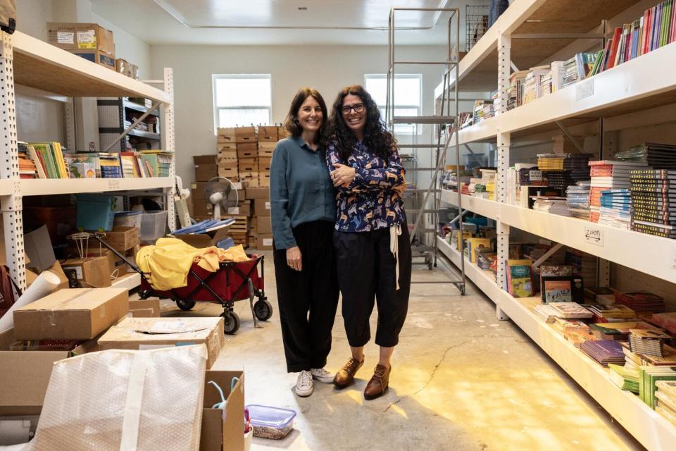 Two women stand between big shelves of books.