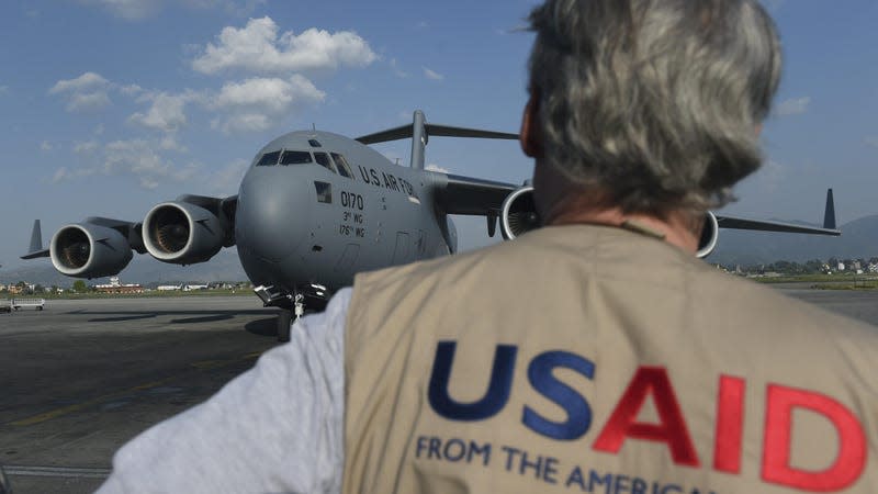 A man wearing a khaki vest with "USAID" on the back in red and blue and shown.