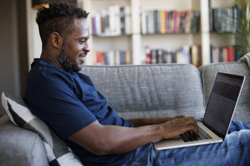 A person is seated on a couch, using a laptop and smiling. There is a bookshelf in the background