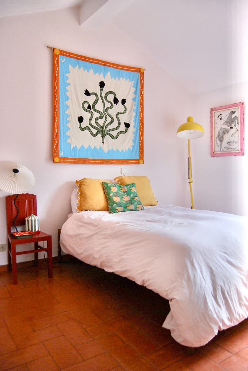 Tapestry art hung above bed centered between wood chair and yellow floor lamp in bedroom.