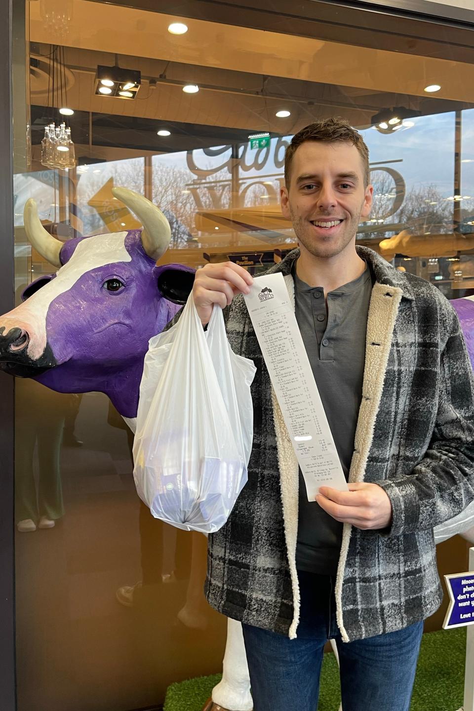 Man holding a long receipt, standing by a statue of a cow outside a cafe