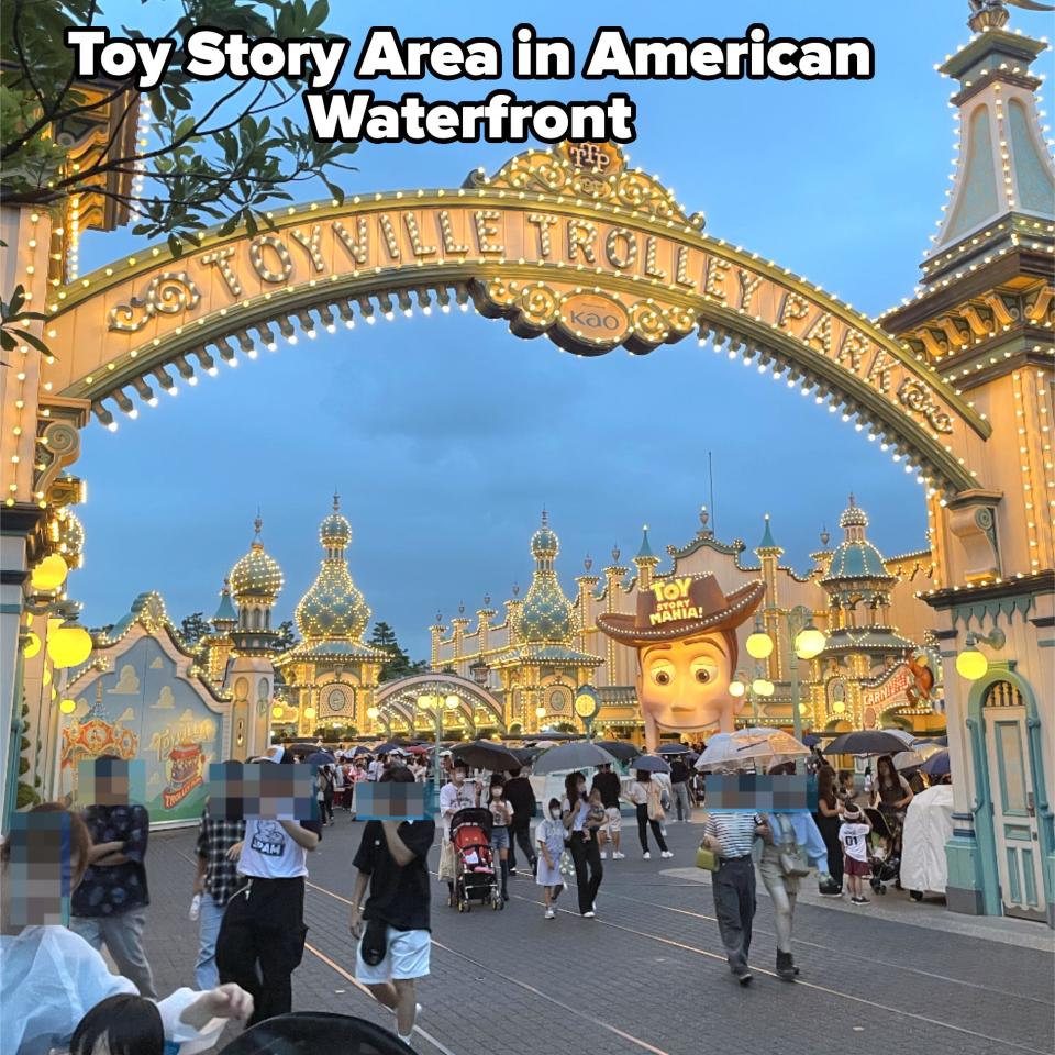 People enjoying a visit to Toyville Trolley Park, passing through its illuminated archway with various whimsical buildings in the background