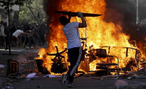 An anti-government protester adds an object to burning chairs and benches amid a march by students and union members in Santiago, Chile, Monday, Oct. 21, 2019. Protesters defied an emergency decree and confronted police in Chile’s capital on Monday, continuing disturbances that have left at least 11 dead and led the president to say the country is “at war.” (AP Photo/Miguel Arenas)